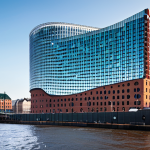 A majestic, iconic architectural masterpiece, the Elbphilharmonie in Hamburg, showcasing its striking contrast between the historic, red-brick base and the futuristic, wave-like glass superstructure. The intricate glass facade dynamically reflects the sky and the Elbe River, creating a sense of movement and grandeur. The building stands prominently against a clear sky, with the bustling Hamburg port and distant city skyline visible in the background. Professional architectural photography, high resolution, sharp focus, detailed textures, natural lighting, safe for work, appropriate content, fully clothed, professional.